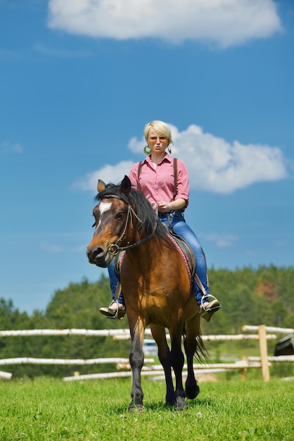 Mujer feliz con gafas de sol sentada en un animal de granja de caballos al aire libre con el cielo azul en el fondo