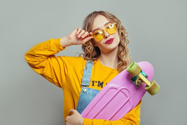 Mujer feliz con gafas de sol sosteniendo una patineta rosa sobre un fondo blanco