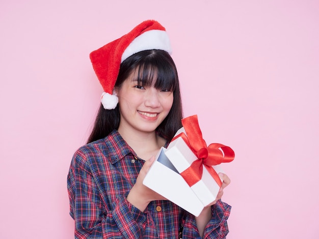 Mujer feliz con gorro de Papá Noel abriendo caja de regalo de Navidad en pared rosa