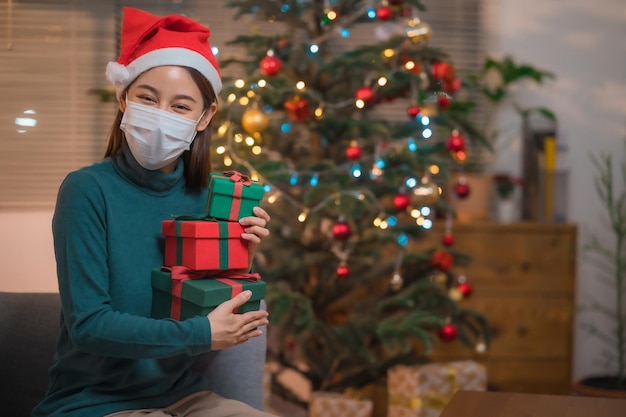 Mujer feliz con gorro de Papá Noel y máscara médica protectora sonriendo mientras sostiene la caja de regalo durante la celebración de Navidad y año nuevo solo durante el brote del virus corona, auto cuarentena.