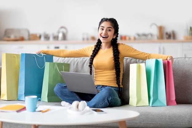 Mujer feliz de grandes ventas con laptop sentada entre bolsas de compras en casa