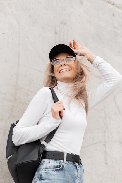 Una mujer feliz y hermosa con una linda sonrisa vestida de forma casual con una gorra negra, un suéter blanco, una mochila y gafas, camina y disfruta en la calle cerca de un muro de hormigón