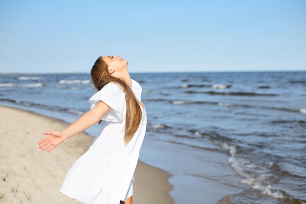 Mujer feliz y hermosa en la playa del océano de pie con un vestido blanco de verano, con los brazos abiertos.