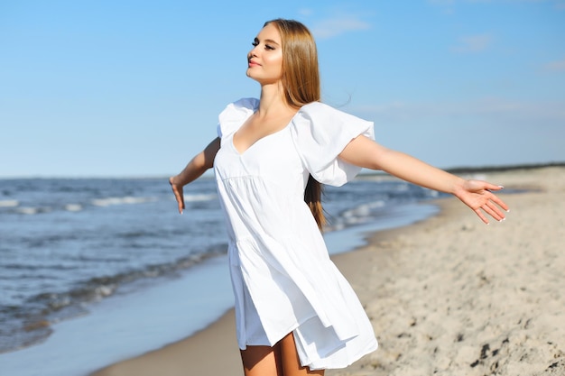 Mujer feliz y hermosa en la playa del océano de pie en un vestido blanco de verano, brazos abiertos.