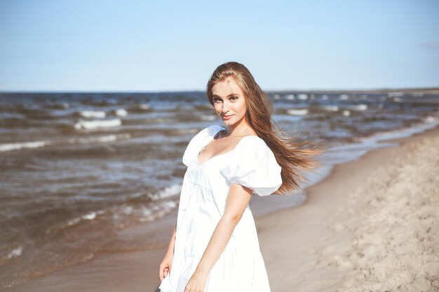 Mujer feliz y hermosa en la playa del océano de pie en un vestido blanco de verano.