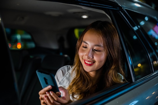 Una mujer feliz y hermosa que envía mensajes de texto con un teléfono inteligente sentada en el asiento trasero de su auto en una mujer de negocios asiática urbana que viaja desde la oficina en el asiento trasero de un taxi con un teléfono móvil en la carretera en la ciudad por la noche después del trabajo tardío