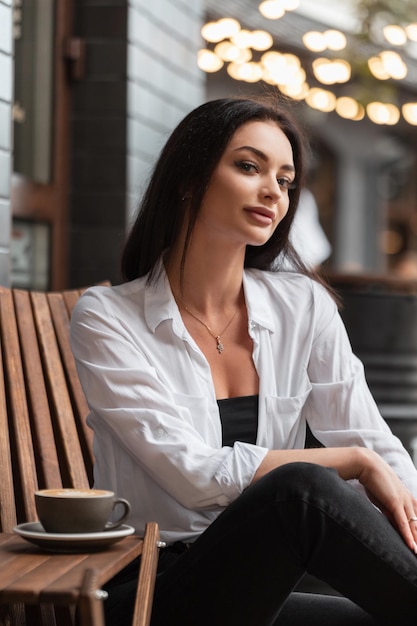 Una mujer feliz y hermosa con ropa informal de moda y una camisa blanca se sienta en un café urbano de verano y bebe café