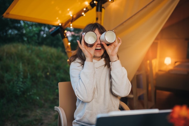 Mujer feliz independiente que usa una computadora portátil y toma café en una acogedora carpa glamping Tienda de campaña de lujo para vacaciones de verano al aire libre y vacaciones Concepto de estilo de vida