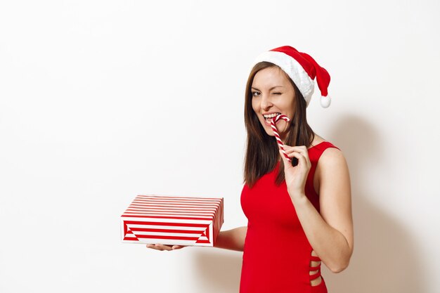 Mujer feliz joven bastante caucásica con sonrisa encantadora vestida con vestido rojo y sombrero de Navidad con caja de regalo y fondo blanco de piruleta. Niña de Santa con presente aislado. Vacaciones de año nuevo 2018.