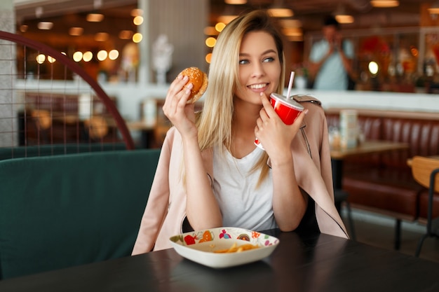 Mujer feliz joven divertida con una hamburguesa y cola comiendo comida rápida en un café