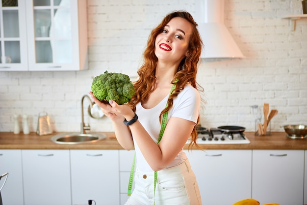 Mujer feliz joven que sostiene el bróculi en la cocina hermosa con los ingredientes frescos verdes dentro. Comida sana y concepto de dieta. Perdiendo peso