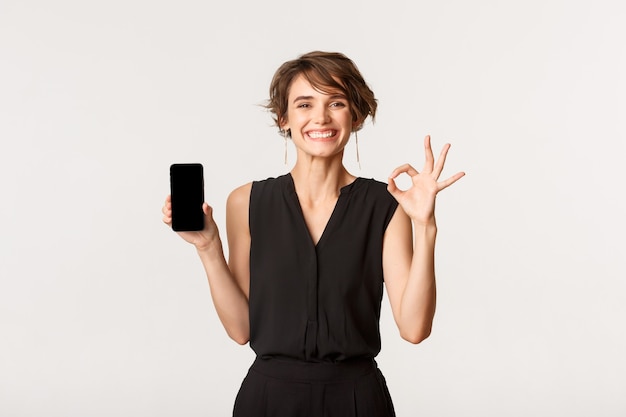 Mujer feliz joven satisfecha que muestra la pantalla del teléfono móvil y un gesto bien, garantizar o recomendar algo, de pie en blanco.