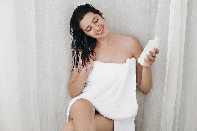 Foto mujer feliz joven en toalla blanca sosteniendo acondicionador para el cabello y sonriendo en el baño cuidado de la piel y el cabello mujer sexy delgada con piel natural disfrutando de spa y bienestar relajante espacio de copia