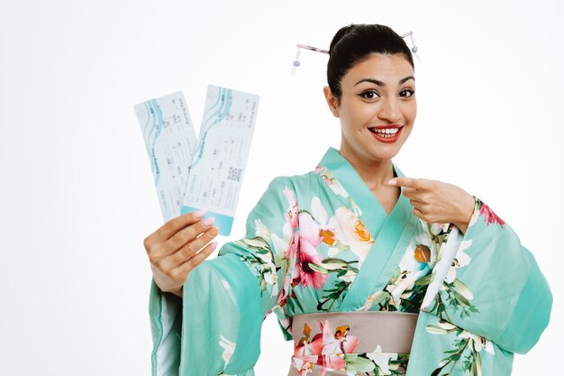 Mujer feliz en kimono japonés tradicional sosteniendo boletos de avión apuntando con el dedo índice a ellos en blanco