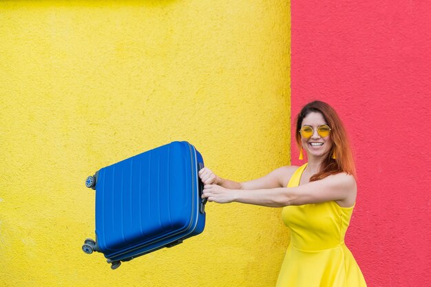 Foto mujer feliz en un látigo sobre un fondo amarillo rojo alegremente agita una maleta antes de viajar hermosa chica en gafas de sol con una bolsa azul en sus manos equipaje para las vacaciones de verano