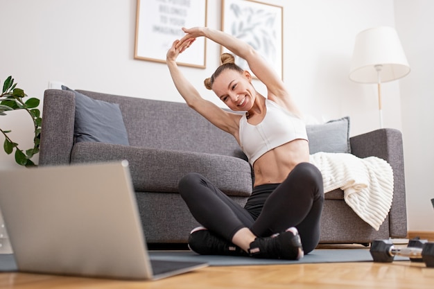 Mujer feliz levantando los brazos y estirando el cuerpo mientras está sentada con las piernas cruzadas cerca de la computadora portátil y haciendo ejercicio en línea en casa durante la pandemia