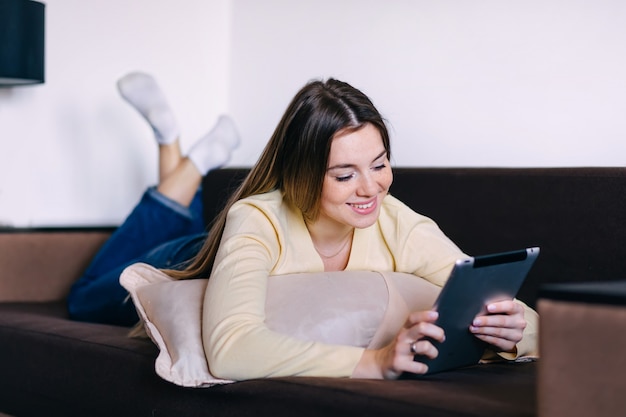 Mujer feliz leyendo ebook en lector digital mientras está acostado en el cómodo sofá