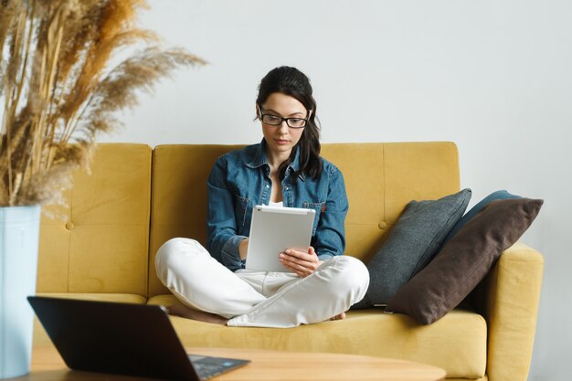 Mujer feliz leyendo un libro en un lector de libros electrónicos sentado en un sofá en casa