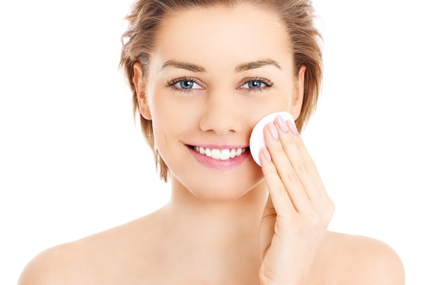 Una mujer feliz limpiando su rostro con algodones sobre fondo blanco.