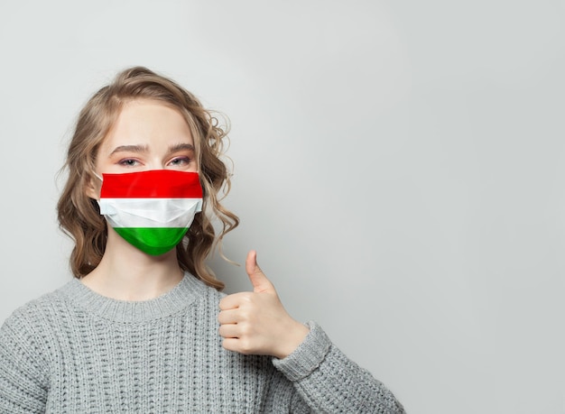 Mujer feliz con máscara facial sosteniendo el pulgar hacia arriba con la bandera de Hungría en el fondo Epidemia de gripe y concepto de protección contra el virus