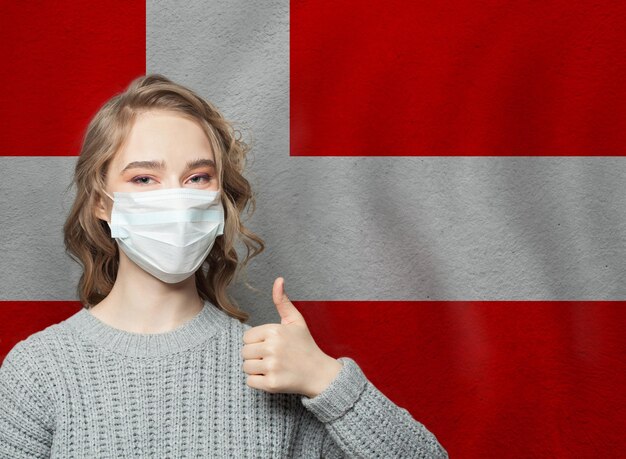 Foto mujer feliz con máscara facial sosteniendo el pulgar en la bandera nacional dinamarca fondo epidemia de gripe y concepto de protección contra el virus