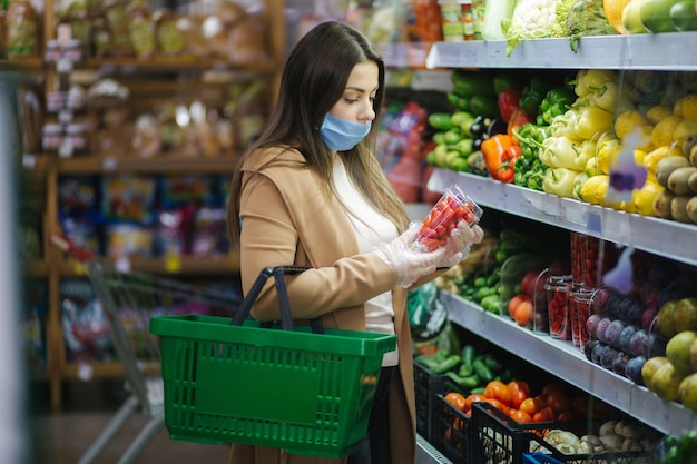 Mujer feliz en mascarilla tomando tomates cherry frescos mientras está de pie en la tienda de comestibles en el supermercado