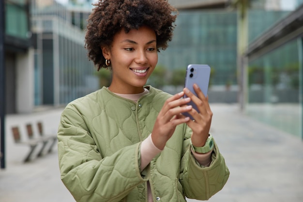 Mujer feliz mira la pantalla del teléfono móvil utiliza un dispositivo moderno para las poses de video chat al aire libre contra el fondo borroso lleva una chaqueta tiene paseos de buen humor en un entorno urbano. Tecnología y estilo de vida