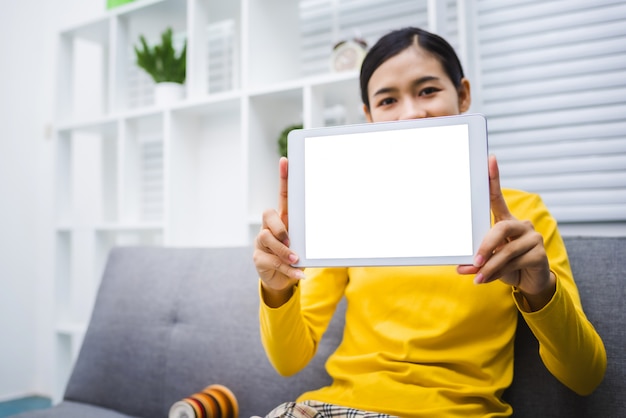 La mujer feliz de la muchacha asiática joven que se sienta en el sofá muestra la tableta blanca de la pantalla.