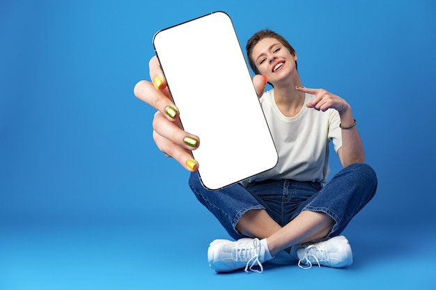 Mujer feliz muestra la pantalla del teléfono inteligente en blanco contra el fondo azul.