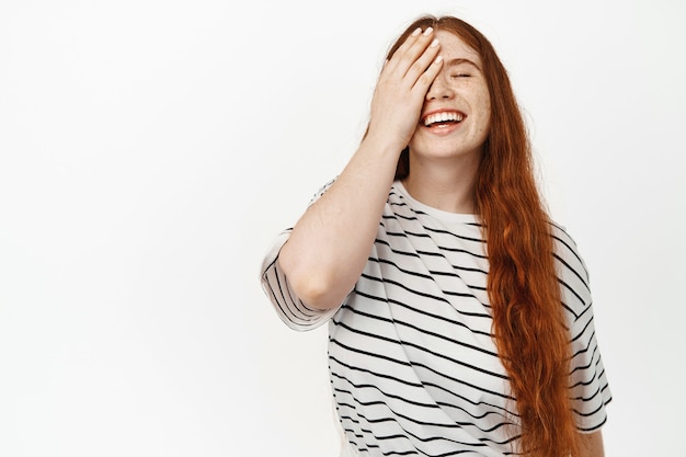 Mujer feliz natural con cabello largo rojo jengibre, riendo despreocupado, con la mitad de la cara con la mano y sonriendo alegre, de pie en camiseta, expresión facial positiva en blanco