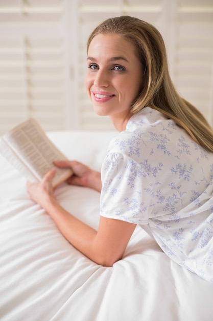 Mujer feliz natural que miente en la cama que sostiene el libro