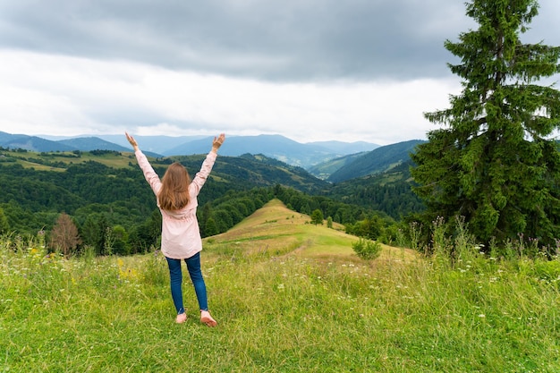 Mujer feliz parada en una colina verde y mirando las montañas con las manos en alto. paisaje de la naturaleza