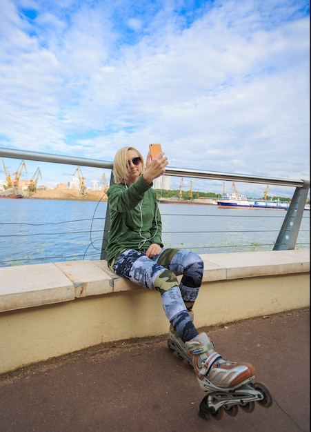 Foto mujer feliz en patines sentado y haciendo foto selfie en teléfono inteligente en riverside