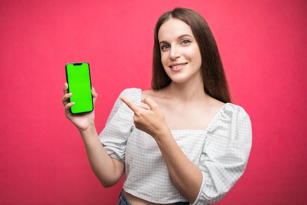 Mujer feliz de pie y sosteniendo el teléfono inteligente y apuntando al teléfono con fondo rosa. Teléfono con pantalla verde