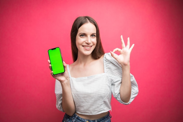 Mujer feliz de pie y sosteniendo el teléfono inteligente y gestando bien sobre fondo rosa. Teléfono de pantalla verde