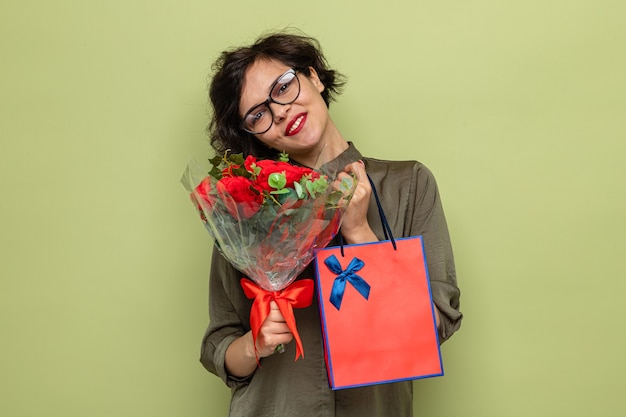 Mujer feliz y positiva con el pelo corto con ramo de flores y bolsa de papel con regalos sonriendo alegremente celebrando el día internacional de la mujer