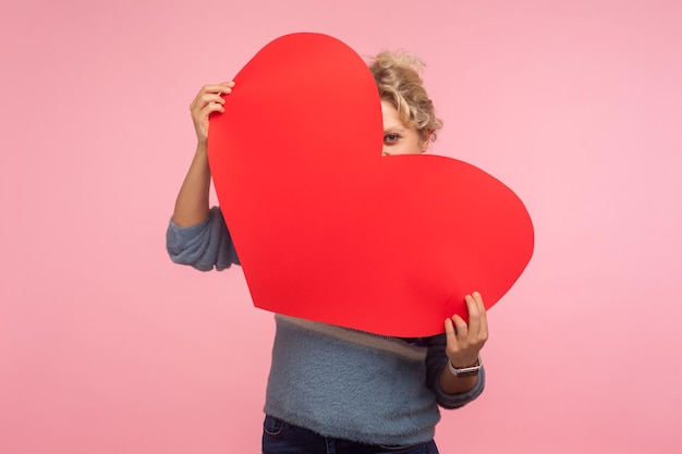 Mujer feliz positiva con el pelo rizado escondiendo la cara detrás de un gran corazón rojo y mirando a la cámara con curiosos ojos indiscretos expresando te amo sintiendo afecto cariño foto de estudio fondo rosa