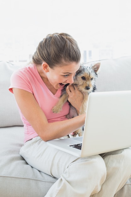 Mujer feliz que sostiene su yorkshire terrier en el sofá usando la computadora portátil