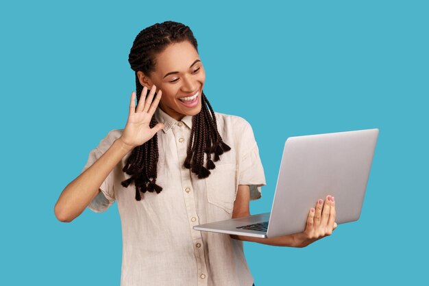 Mujer feliz con rastas negras conversando en línea, saludando con la mano y saludando, mirando la cámara web de la laptop, usando camisa blanca. Disparo de estudio interior aislado sobre fondo azul.