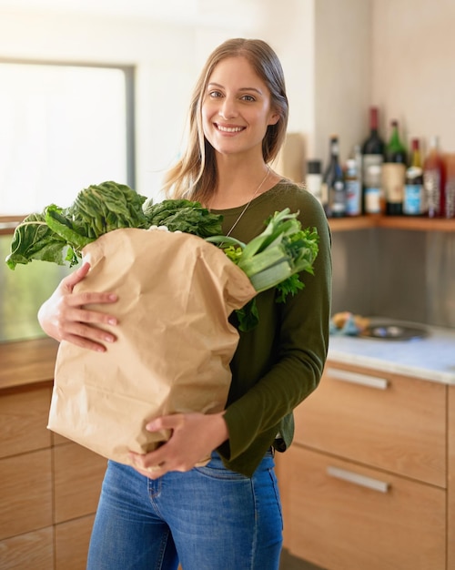 Mujer feliz y retrato con verduras en la cocina para comprar comestibles cocina y alimentación saludables sonrisa de persona femenina y bolsa de ingredientes con espinaca o lechuga para nutrición y dieta vegana