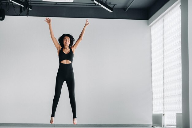 Una mujer feliz en ropa deportiva negra hace un salto sobre un fondo blanco Chica saltando levanta las manos en el gimnasio