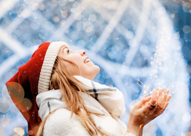 Mujer feliz con ropa de invierno sosteniendo la luz de Navidad y sonriendo en el frío día de invierno nevando