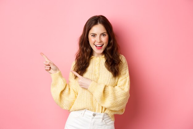 Mujer feliz segura de sí misma con peinado rizado, señalando con el dedo a un lado, mostrando promo en el copyspace izquierdo, sonriendo emocionada, de pie sobre una pared rosa.