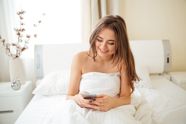 Mujer feliz sentada en la cama debajo de la manta y con smartphone en casa