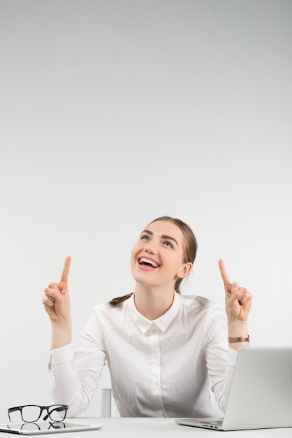 La mujer feliz se sienta al lado de una computadora portátil y la risa mira hacia arriba apuntando los dedos hacia arriba en sus dos manos. - Imagen vertical