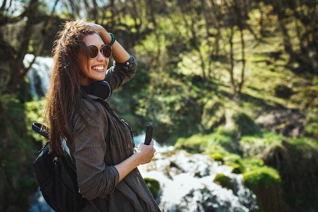 Una mujer feliz, con un smartphone en la mano y auriculares alrededor del cuello, disfruta del paisaje en las montañas mirando la hermosa cascada al aire libre.