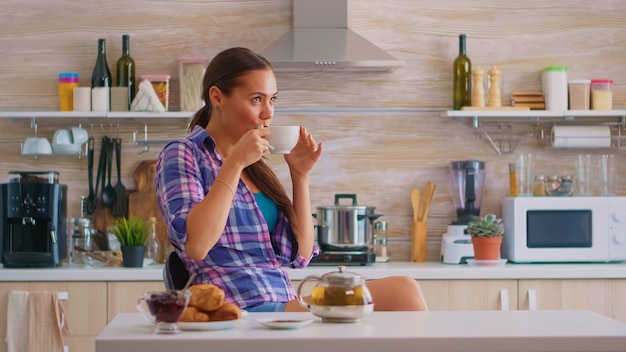 Mujer feliz soñadora tener una gran mañana bebiendo té verde caliente. Señora alegre sentada en la cocina durante el desayuno relajándose con un sabroso té de hierbas naturales de la taza de té blanca.