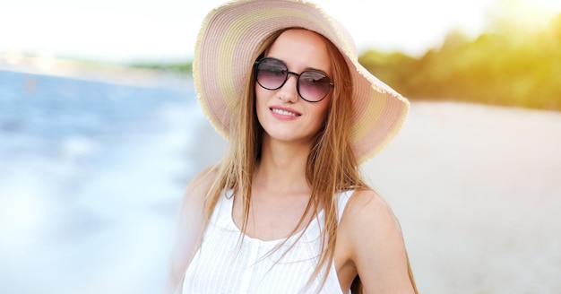 Mujer feliz sonriente en felicidad libre en la playa del océano de pie con sombrero, gafas de sol y flores blancas. Retrato de una modelo femenina multicultural en vestido blanco de verano disfrutando de la naturaleza