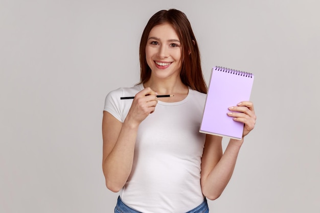 Mujer feliz sonriente positiva apuntando al cuaderno de papel con espacio en blanco para el área de publicidad de texto comercial con camiseta blanca Foto de estudio interior aislada en fondo gris