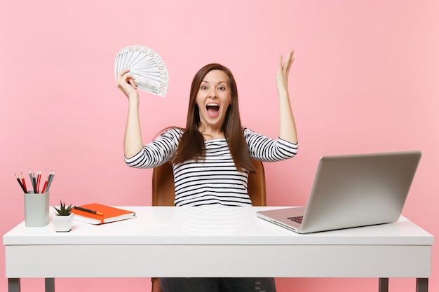 Mujer feliz sorprendida extendiendo las manos sosteniendo un montón de dólares, dinero en efectivo trabajando en la oficina en el escritorio blanco con computadora portátil pc